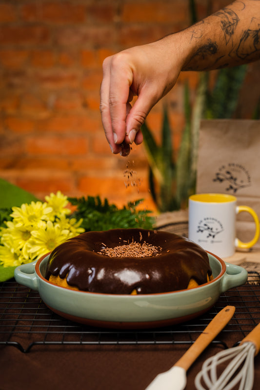 Pound carrot cake cover with a lot brigadeiro, sitting at green dish, flower on the background and mug white and yellow. whisk and spatula decorating. A hand sprinkling chocolate sprinkles 