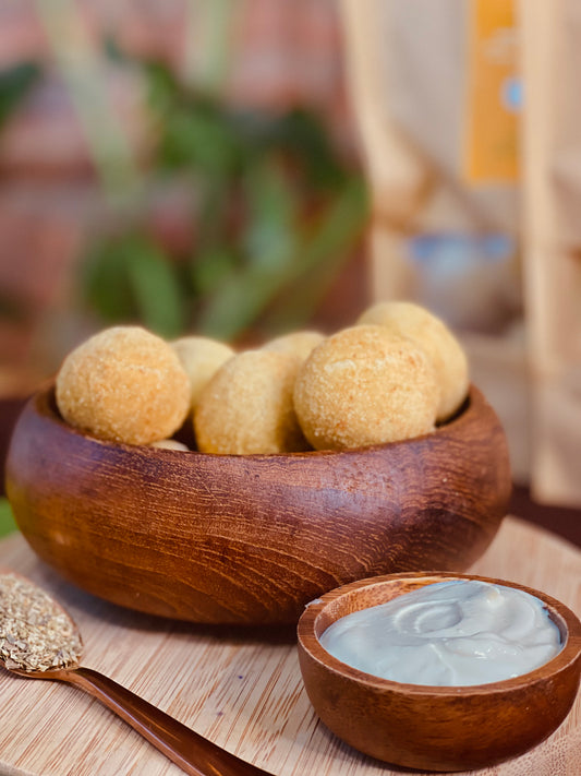 Vegan creamy corn croquet, inside of a wood bowl, next to tiny wood bowl with cream cheese, next to a copper spoon with oregano on top of wood board, with green plants on the background 