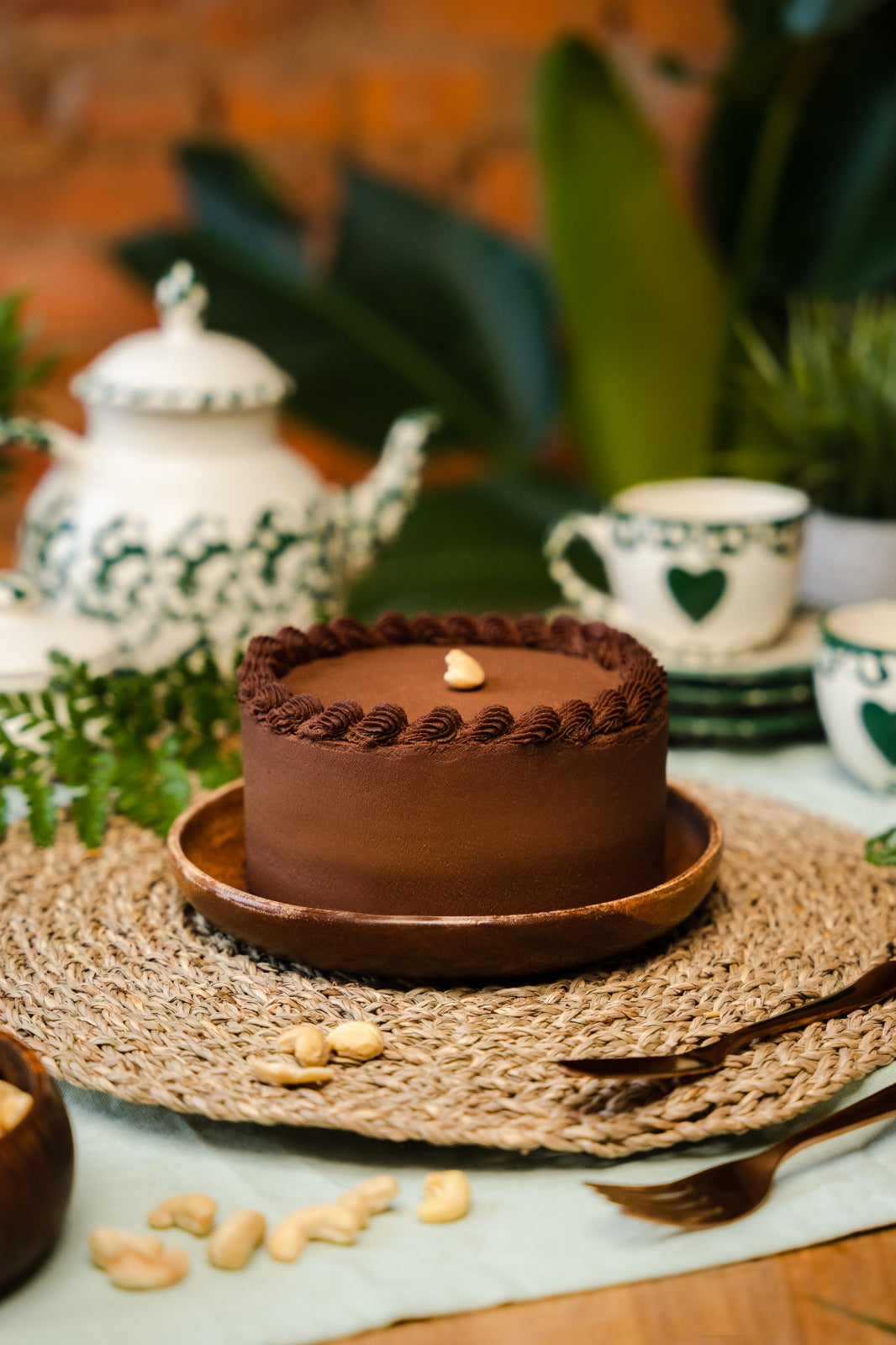 A chocolate cake display in a wood plate, with some cashew around, fork, knife, mugs and teapot for decoration