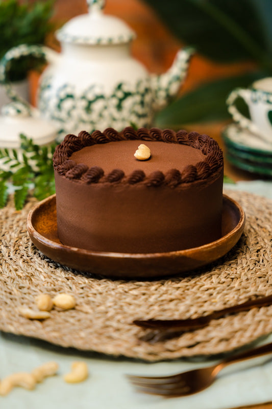 A chocolate cake display in a wood plate, with some cashew around, fork, knife, mugs and teapot for decoration